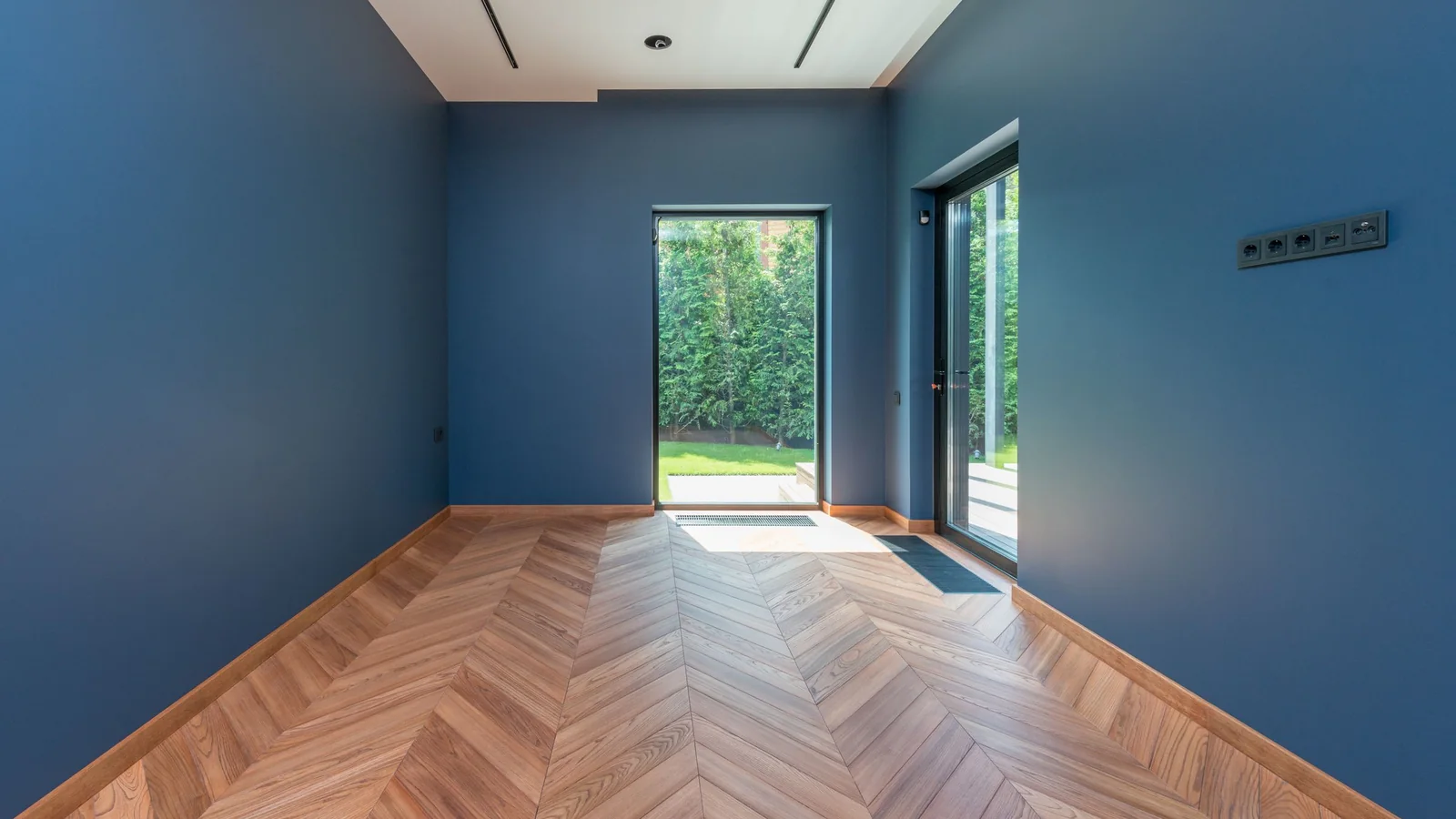 Pièce vide aux murs bleus et parquet en chevrons, porte ouverte sur un jardin